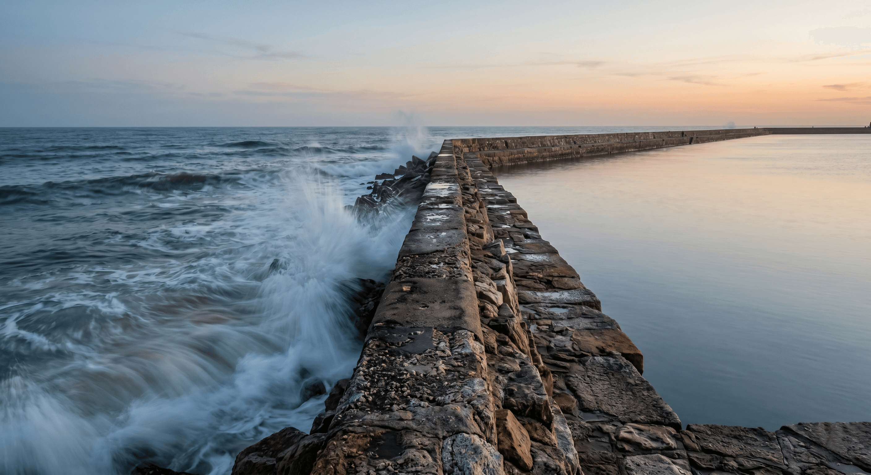 A long-exposure, artistic shot of a stone breakwater or sea wall extending into a calm ocean at dusk. On one side of the wall, the water is turbulent; on the other, it is perfectly still.